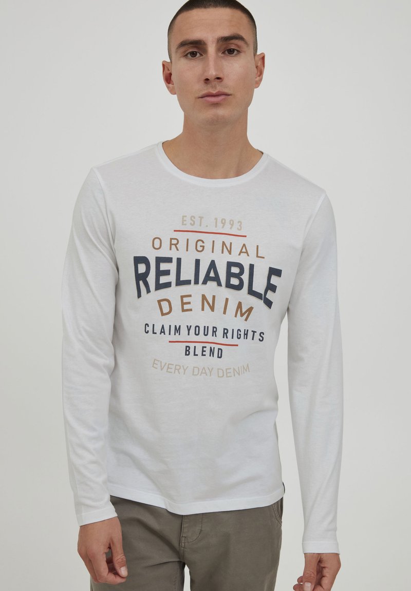 Hvit langermet T-skjorte med rund hals, med et flerfarget tekstgrafikk: "Original Reliable Denim" og tilleggsetninger i forskjellige farger.