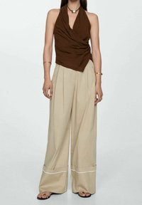 Top corto marrone con design drappeggiato abbinato a pantaloni a gamba larga beige con un dettaglio in bianco sul fondo. Inclusi semplici accessori in gioielleria.