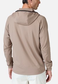 Tan letvægshætteklædning med en tætsiddende silhuet, justerbar hætte og sømløse syninger, båret over hvide bukser.