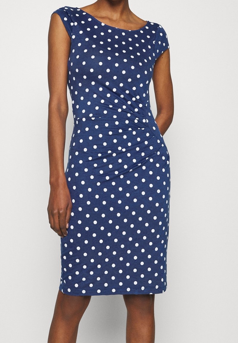 Femme portant une robe bleu marine ajustée à pois blancs avec des manches courtes, debout devant un fond clair uni.