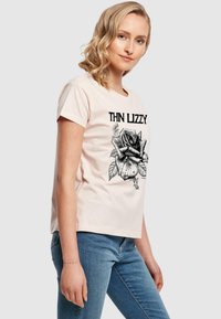 Šviesiai rožinė medvilninė marškinė su juodos rožės grafiką ir užrašu "THIN LIZZY." Trumpomis rankovėmis ir laisvu siluetu.