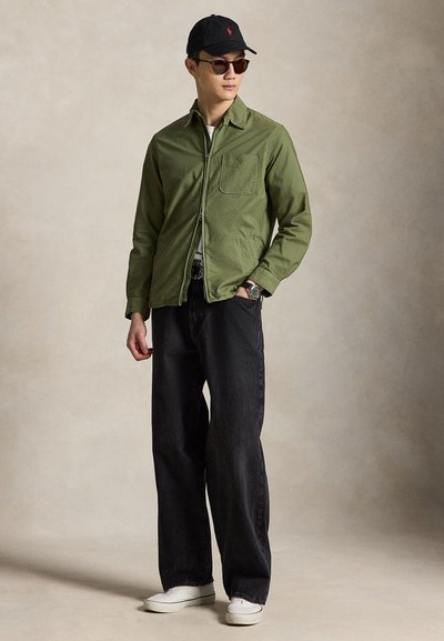 Mann, der schwarze Mütze und Sonnenbrille trägt, olivgrüne Jacke mit Reißverschluss, schwarze weit geschnittene Jeans, weiße Sneaker, steht mit einer Hand in der Tasche.