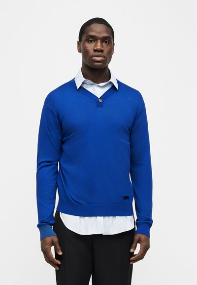 Νεαρός άνδρας που στέκεται, φορώντας ένα φωτεινό μπλε πουλόβερ με V λαιμόκοψη πάνω από ένα ανοιχτόχρωμο ριγέ πουκάμισο με γιακά και μαύρο παντελόνι μπροστά σε λευκό φόντο.