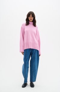 Maglione oversize rosa con collo alto in morbido tessuto a maglia, abbinato a jeans blu larghi con cuciture bianche e scarpe basse nere.