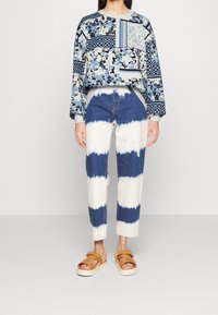Felpa con motivo in tonalità blu; pantaloni in denim con strisce tie-dye blu e bianche; sandali con piattaforma color beige con dettaglio della fibbia.