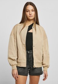 Giacca bomber beige con una superficie liscia e lucida, dotata di chiusura frontale a zip e polsini elastici. Indossata sopra una maglietta nera e pantaloni corti.