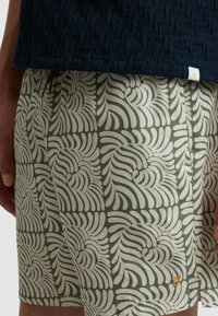 Zwemshorts met een lichtgroen en crèmekleurig wervelend bladerenpatroon, gemaakt van een gladde stof. Inclusief een klein oranje logo aan de zijkant.