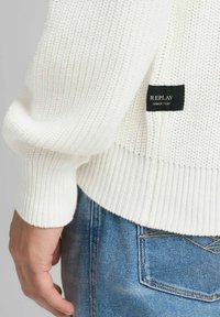 Maglione bianco lavorato a maglia con polsini e orlo a coste, con etichetta nera recante la scritta "REPLAY SINCE 1981". Abbinato a jeans di denim blu.