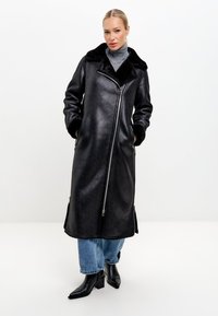 Černý kabát z umělé kůže s velkým koženým límcem, dlouhými rukávy, předním zipem a bočními kapsami. Nositelný přes světle modré džíny a černé boty.
