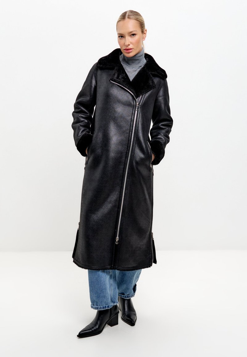 Černý kabát z umělé kůže s velkým koženým límcem, dlouhými rukávy, předním zipem a bočními kapsami. Nositelný přes světle modré džíny a černé boty.