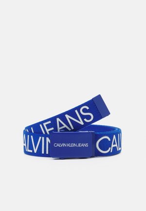 Niebieski materiałowy pasek z białym napisem „CALVIN KLEIN JEANS” oraz dopasowaną niebieską klamrą z tym samym białym napisem na jednolitym tle.