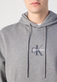 Pilkas gobtuvas su raišteliais, su siuvinėtu "Calvin Klein" logotipu ir tekstūruota medžiaga. Paprastas dizainas, be raštų ar akcentų.