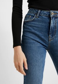 Pantalones vaqueros de tiro alto en denim azul con una textura suave, diseño de cinco bolsillos y costuras en contraste, combinados con una blusa negra de mangas largas.