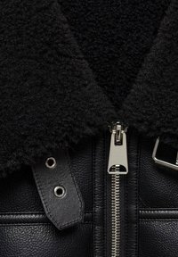 Giacca nera in pelle con colletto peloso nero, con una cerniera argento, chiusure a scatto e una superficie testurizzata.