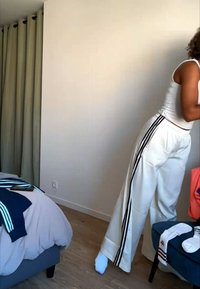 Personne portant un pantalon blanc à rayures noires et un haut blanc sans manches se penche près d'un lit et d'une chaise dans une pièce peu meublée.