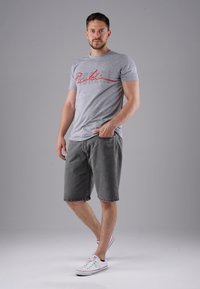 Man stands with one hand in pocket wearing grey graphic t-shirt, dark grey knee-length shorts, and white sneakers against plain grey background.