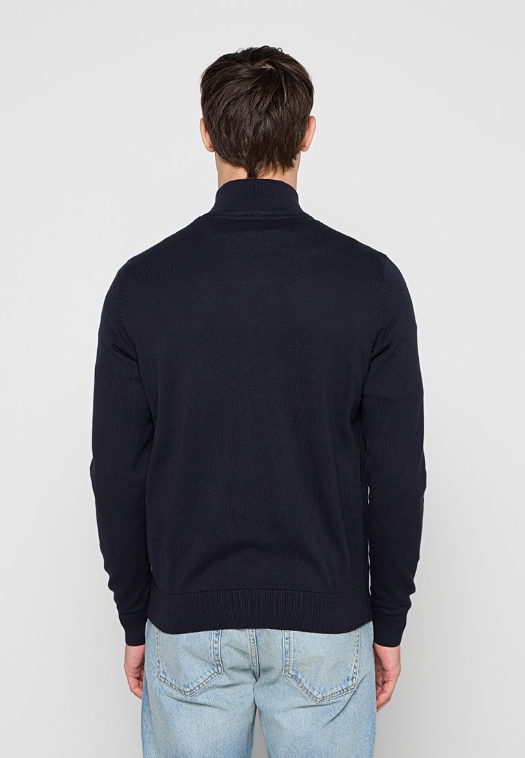 Mann mit kurzen dunklen Haaren, der einen dunkelblauen Pullover mit langen Ärmeln und hellblaue Jeans trägt, mit dem Rücken zu einem einfarbigen hellgrauen Hintergrund.