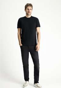 Camiseta de algodón negra, de cuello redondo, combinada con unos vaqueros negros ajustados. El modelo está de pie, vistiendo zapatillas blancas, sobre un fondo neutro.