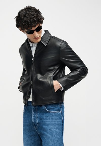 Homme aux cheveux bouclés portant des lunettes de soleil noires, une veste en cuir noire, une chemise rayée et un jean bleu, les mains dans les poches de la veste.