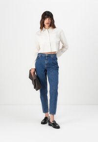 Camisa blanca corta con botones y dos bolsillos frontales, combinada con vaqueros azules de cintura alta y mocasines negros, llevando un bolso de mano gris.
