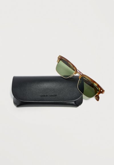 Γυαλιά ηλίου με σκελετό από χελώνα και πράσινους φακούς, τοποθετημένα πάνω σε μαύρη δερμάτινη θήκη Giorgio Armani σε λευκό φόντο.