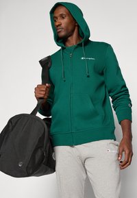 Groene ritshoodie met trekkoord, voorzien van een wit logo. Gecombineerd met grijze joggingbroek en een zwarte sporttas, wat een eenvoudige atletische stijl laat zien.