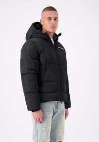 Zwarte pufferjack met een ritssluiting aan de voorkant, een vaste capuchon en een horizontaal gestikt patroon. Voorzien van zijzakken en minimale branding op de borst.