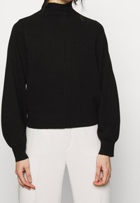 Svart höghalssweater i stickad kvalitet med långa ballongsärmar och ribbad fåll. Slät struktur med sid sömsdetalj. Stylingen kompletteras med vita byxor.