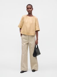 Femme portant un chemisier ample beige brodé, un pantalon clair à jambes larges, des chaussures noires, tenant un sac à main noir sur un fond uni.