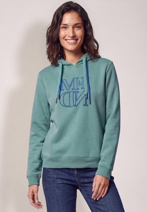 Femme souriante aux cheveux foncés mi-longs porte un sweat à capuche bleu sarcelle avec des cordons bleus et des inscriptions, assorti à un jean bleu foncé.