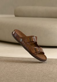 Sandale en cuir marron à enfiler avec deux boucles ajustables et semelle texturée, présentée flottant au-dessus d'un tapis beige et d'un canapé en arrière-plan.