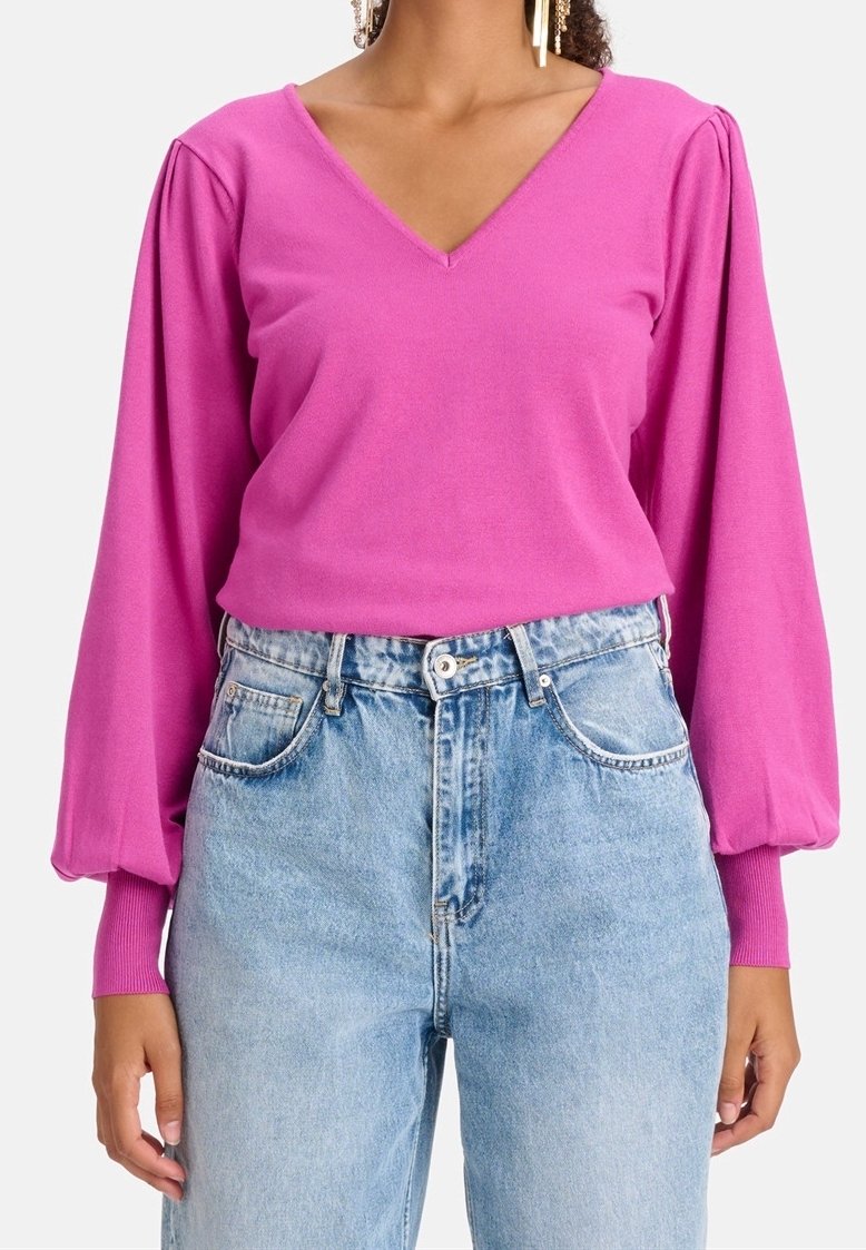 Femme portant un haut rose vif à manches longues avec un col en V et des manches bouffantes, ainsi qu'un jean taille haute bleu clair.