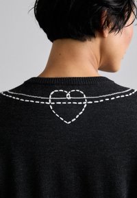 Fekete kötött pulóver, amelyen fehér hímzett szívmotívum található, valamint egy vízszintes vonal a háton a nyakrészen.
