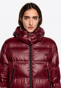 Bordowa kurtka puchowa z wysokim kołnierzem, zamkiem błyskawicznym, dwoma bocznymi zamkami oraz błyszczącym wykończeniem. Posiada regulowane sznurki w kapturze.