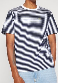 Man wearing navy and white horizontal striped short-sleeve shirt with small green crocodile logo on chest and light blue jeans.