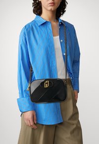 Personne vêtue d'une chemise rayée bleu et blanc, de pantalons beiges et d'un sac à bandoulière noir avec un fermoir en or, se tenant devant un fond uni.