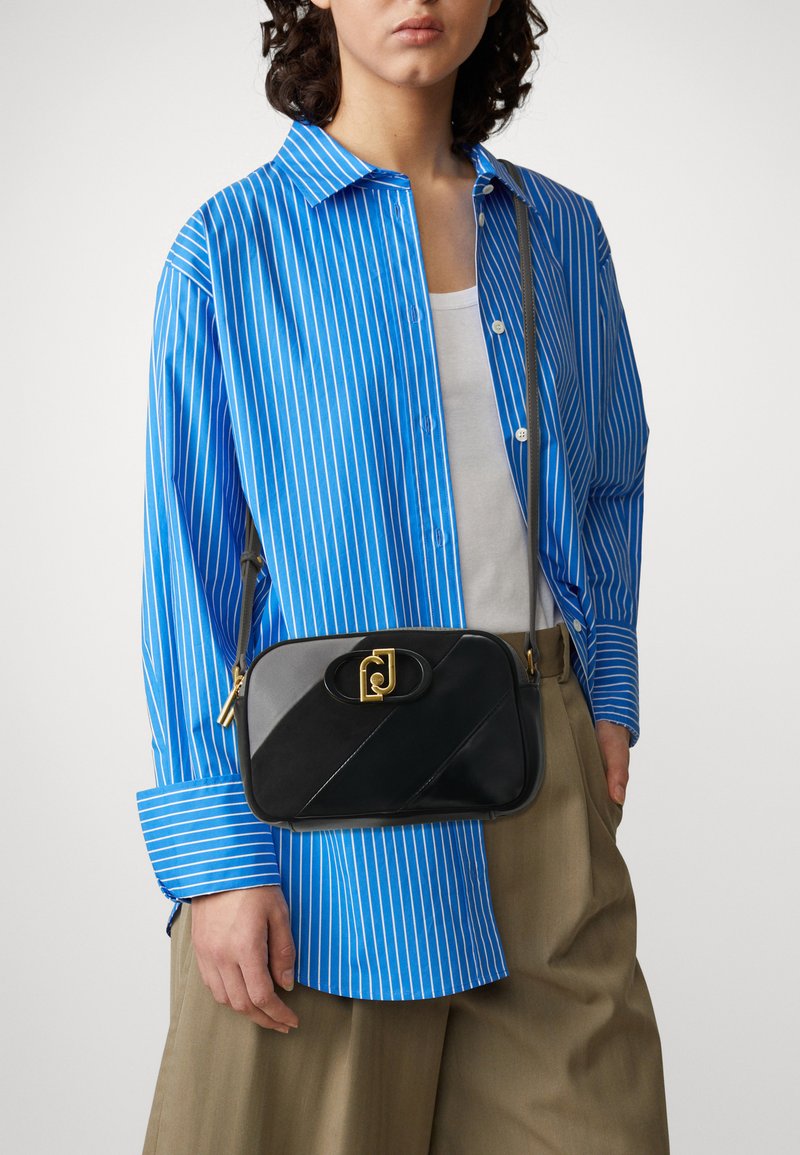 Personne vêtue d'une chemise rayée bleu et blanc, de pantalons beiges et d'un sac à bandoulière noir avec un fermoir en or, se tenant devant un fond uni.