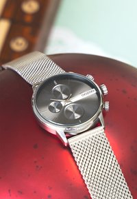 Montre avec un bracelet en maille argentée, un cadran rond noir et argent, trois sous-cadrans et des accents argentés, pr ésentée sur une surface rouge.