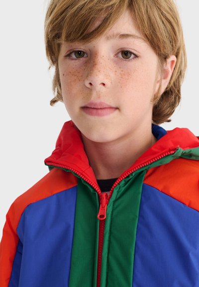 Niño pequeño con pecas y cabello castaño claro, vistiendo una chaqueta colorida con secciones rojas, verdes y azules sobre un fondo liso.