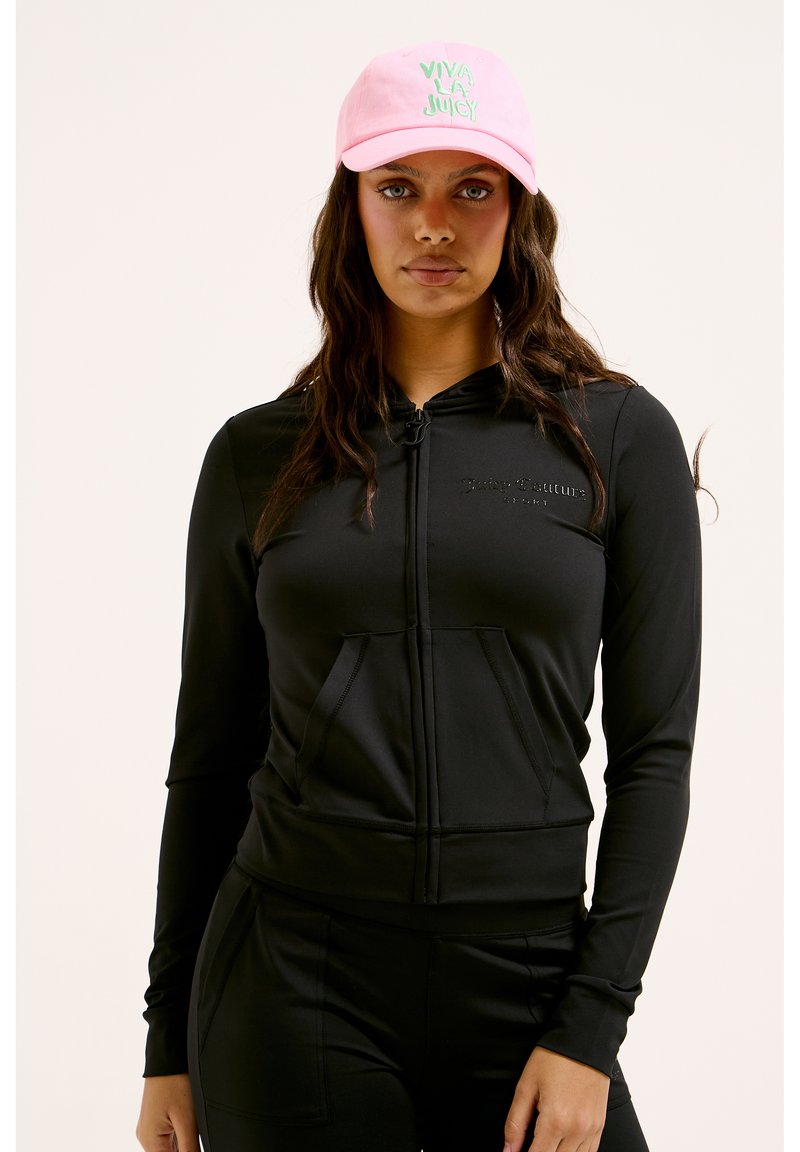 Veste zippée noire à manches longues, en matériau lisse, avec un design à poches doubles. Le modèle porte une casquette rose avec du texte vert.