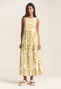 Bela večplastna maxi obleka z rumenimi vzorci listov, okroglo izrezom in lahkotnim tkanjem. Kombinirana s svetlimi platformnimi sandali.