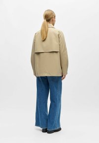 Femme avec queue de cheval blonde portant une veste beige et un jean large bleu, debout de dos sur un fond clair uni.