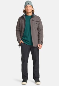 Chaqueta gris aislante con forro interior estampado, camisa azul verdosa, pantalones negros y un gorro azul claro. Calzado casual completa el look.