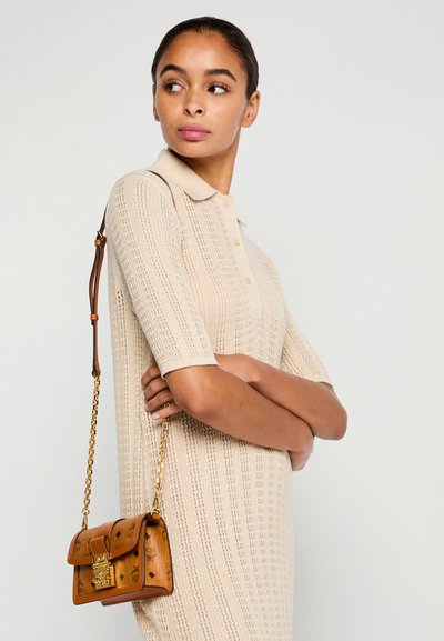 Kvinde med tilbageholdte hår iført en beige strik kjole og en brun læder skuldertaske med guld kæde, ser til siden mod en ensfarvet baggrund.