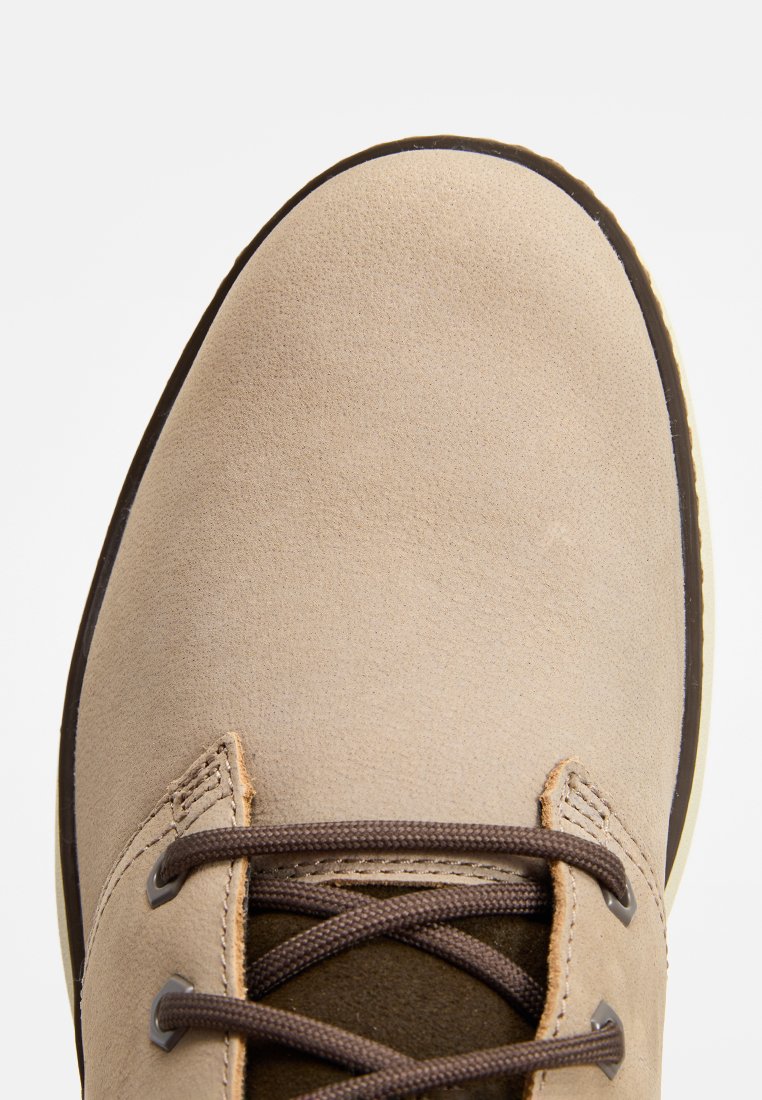 Zapato de ante beige con cordones y detalles marrones, diseño de punta redonda y suela de goma flexible. La superficie tiene una textura suave.