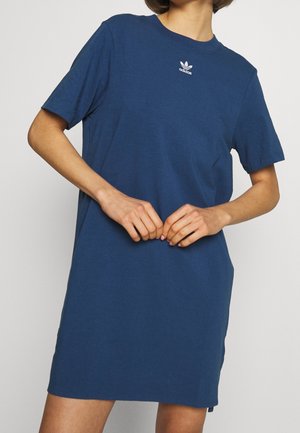 Osoba ubrana w gładką niebieską sukienkę typu t-shirt marki Adidas, ręce złożone na wysokości talii, na jednolitym jasnym tle.