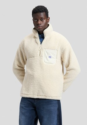 Giovane uomo in piedi indossa un maglione in pile color crema con zip frontale e tasca sul petto, abbinato a jeans blu su uno sfondo chiaro e semplice.