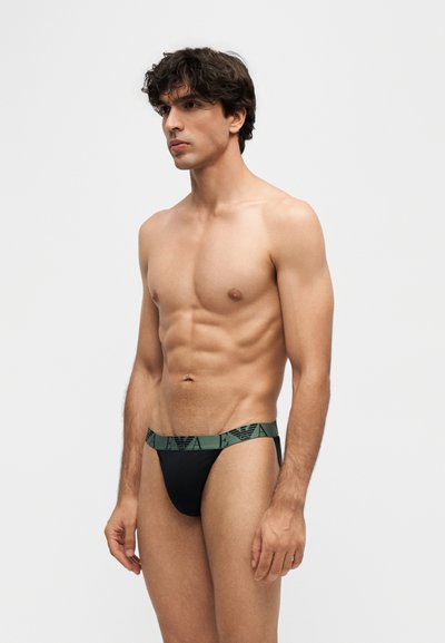 Jeune homme aux muscles toniques portant un slip noir avec une ceinture de marque vert foncé, debout sur un fond clair uni.