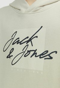 Felpa leggera beige con cappuccio caratterizzata da un logo scritto a mano in nero "Jack & Jones" su uno sfondo a righe, con "SINCE 1990" in caratteri piccoli sotto.