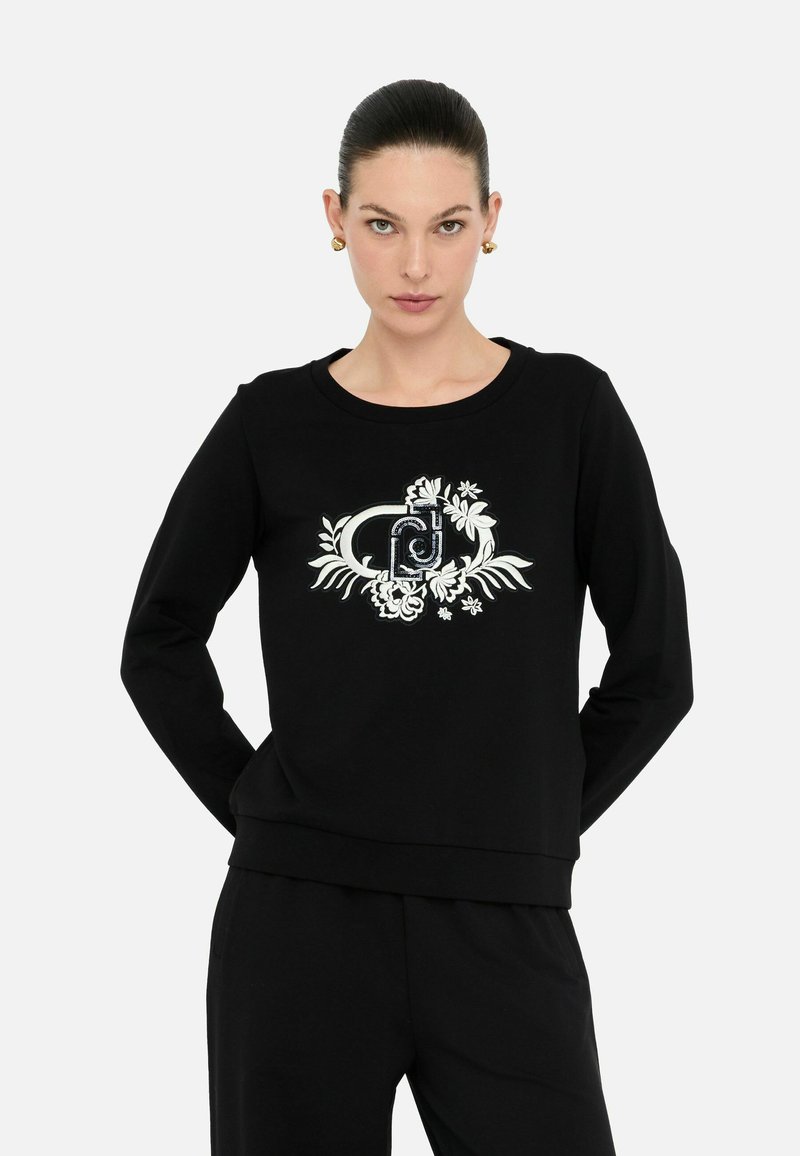 Zwarte sweatshirt met lange mouwen en een ronde hals, met witte bloemenborduursels en een centraal grafisch ontwerp. Glad stoftextuur.